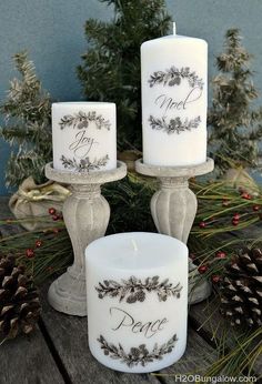 two white candles sitting on top of a table next to pine cones and evergreen branches