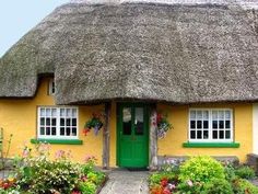 a thatched roof house with flowers and plants around the front door, on a sunny day