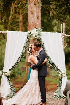 a bride and groom kissing under an arch decorated with greenery at their outdoor wedding ceremony