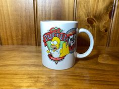 a white coffee mug with the simpsons logo on it sitting on a wooden table next to a wood paneled wall