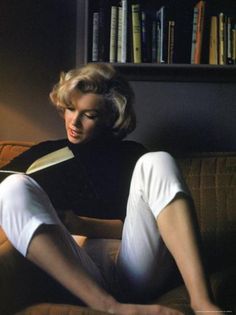 a woman sitting on a couch reading a book with her legs crossed and wearing white pants