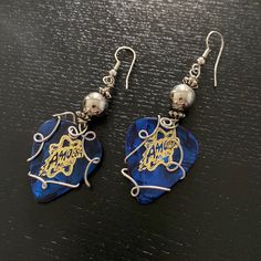 two blue and gold earrings with silver beads on top of a black table next to each other