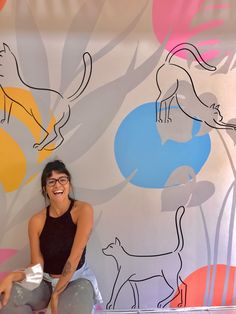 a woman sitting on the ground in front of a wall with cats painted on it