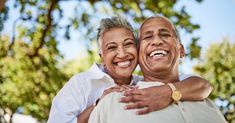 an older couple embracing each other in front of trees and blue sky with sunlight coming through the leaves