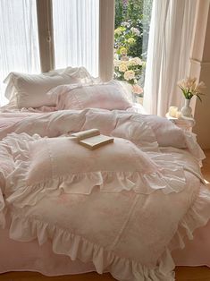 a bed with pink ruffles and pillows in front of a large window that has white curtains