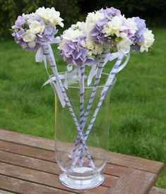 purple and white flowers in a glass vase on a wooden table with grass behind it