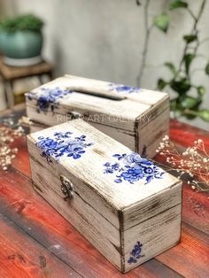 two wooden boxes with blue flowers painted on them sitting on a table next to plants