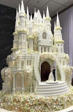 a wedding cake that looks like a castle with white flowers on the side and a bride and groom standing in front