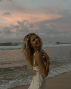 a woman standing on top of a sandy beach next to the ocean at sunset with her hair blowing in the wind