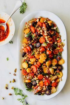 a white platter filled with roasted vegetables and garnished with herbs next to a bowl of sauce