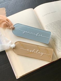 two bookmarks with names on them sitting next to an open book that has a ribbon tied around it