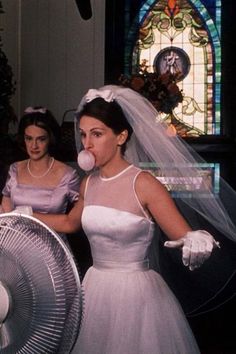 two women dressed in wedding attire standing next to each other and one is blowing a bubble