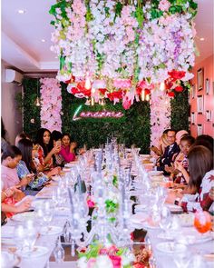 a group of people sitting around a long table with wine glasses on it and flowers hanging from the ceiling