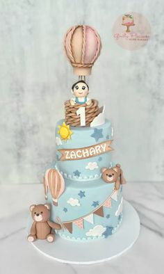 a three tiered blue cake with an air balloon and teddy bear on top that says zachary