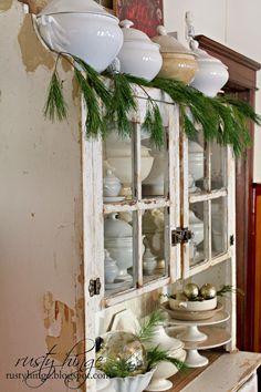 an old china cabinet is decorated with greenery and white dishes on it's glass doors