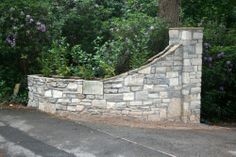 a large stone wall next to a road with trees and bushes in the back ground