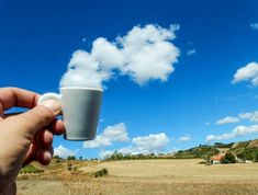 a hand holding a coffee cup in front of a blue sky with white fluffy clouds