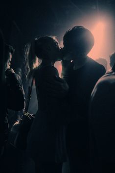 a man and woman kissing in the dark with their arms around each other as people look on