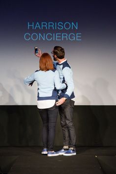 two people standing in front of a stage taking a photo with their cell phones at an event