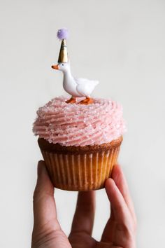 a cupcake with pink frosting and a duck on top is held up by a person's hand