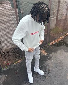 a young man with dreadlocks standing in front of a fence wearing a white sweatshirt and sweatpants