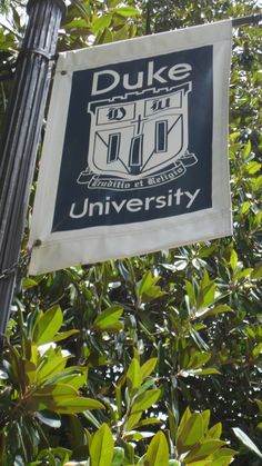 a duke university sign hanging from a metal pole in front of some green bushes and trees