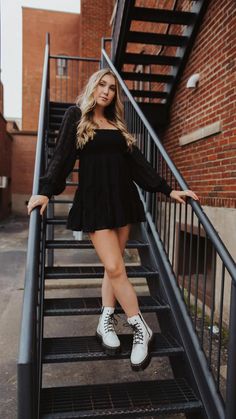 a woman in black dress and white tennis shoes standing on stairs with her hands on the railing