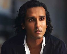 a man with long black hair wearing a white shirt and looking at the camera while sitting down