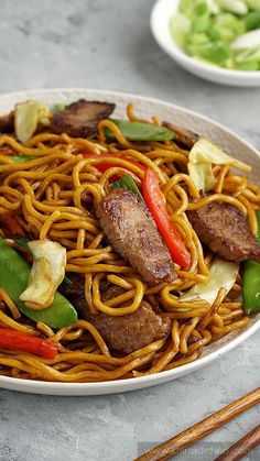 a plate of noodles with meat and vegetables on it next to some chopsticks