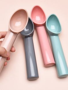 four different colored spoons are shown on a pink surface, with one being held in the other's hand