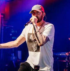 a man on stage holding a microphone in his right hand and wearing a baseball cap