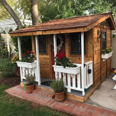 a small wooden shed with potted plants on the front porch and flowers in flower boxes