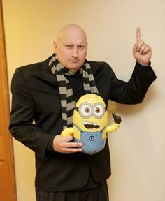 a bald man in a suit and scarf holding a stuffed toy with a minion on it