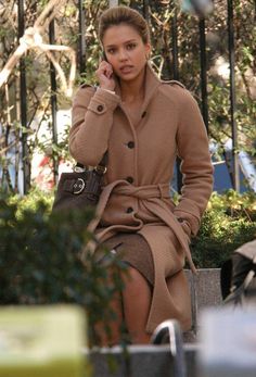 a woman in a trench coat talking on a cell phone while sitting down with her hand up to her ear