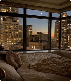 an unmade bed in front of a large window overlooking the city at night time