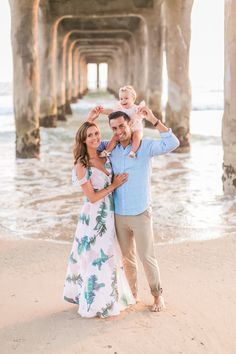 a man and woman holding a baby on the beach under a pier with palm trees