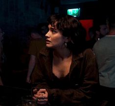 a woman sitting at a table in a dark room with other people around her looking off into the distance