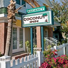 the sign for coconut inn in front of a palm tree and white picket fence with pink flowers