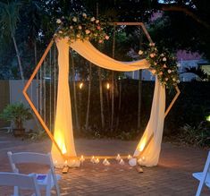 an outdoor wedding setup with white chairs and flowers on the arch, lit up by candles