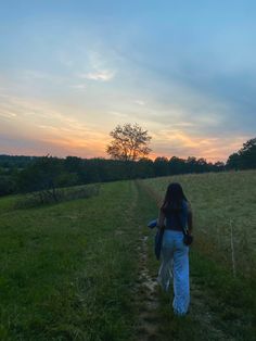 a woman walking down a dirt road in the grass at sunset or dawn with her back to the camera