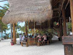 some people are sitting at a table under a thatched roof on the beach with palm trees