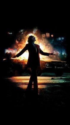 the joker movie poster with his arms outstretched in front of an old car on fire