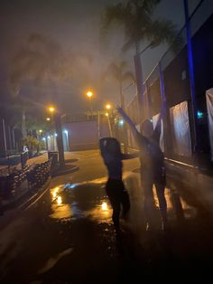 two people standing in the rain at night with their arms up and one person holding an umbrella