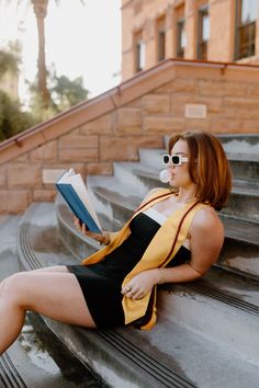 a woman sitting on the steps reading a book with her legs crossed and wearing sunglasses