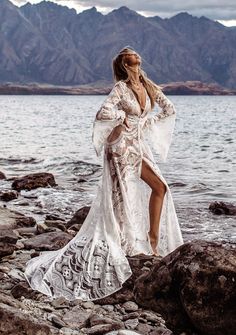 a woman in a white dress standing on rocks by the water with mountains in the background