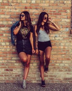 two girls leaning against a brick wall with their hands on their hipss and one holding her hand