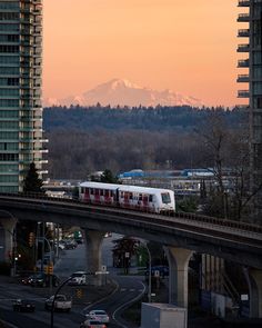 a train traveling over a bridge with tall buildings in the background and a snow capped mountain in the distance