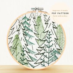 an embroidery pattern with green pine trees in the background on a wooden table next to a white wall