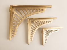 three gold shelf brackets on a white wall, one with holes in the middle and one without