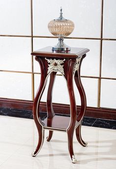 an ornate wooden table with a glass lamp on it's top, in front of a wall